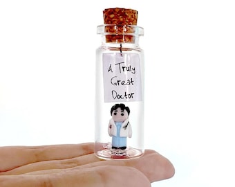 May include: A clear glass bottle with a cork stopper containing a small figurine of a doctor wearing a white coat and blue scrubs. A white paper note inside the bottle features the handwritten black text A Truly Great Doctor.