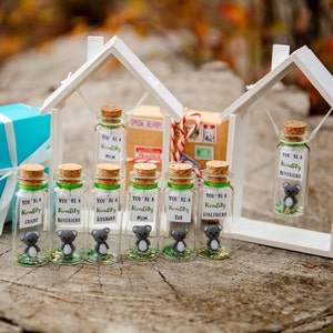 K&ouml;nnte beinhalten: Sieben kleine Glasflaschen mit Korkverschl&uuml;ssen und gr&uuml;nen Deckeln. Jede Flasche enth&auml;lt eine kleine graue Koala-Figur und Glitzer. Die Flaschen sind mit dem Text "You're a Koality" beschriftet, gefolgt von einem anderen Wort auf jeder Flasche, wie "Mama", "Papa", "Ehemann", "Frau", "Freund", "Freundin" und "Freund". Die Flaschen sind in einer Reihe auf einer Holzoberfl&auml;che angeordnet.