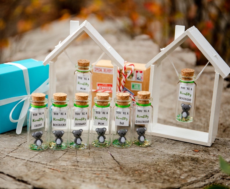 K&ouml;nnte beinhalten: Sieben kleine Glasflaschen mit Korkverschl&uuml;ssen und gr&uuml;nen Deckeln. Jede Flasche enth&auml;lt eine kleine graue Koala-Figur und Glitzer. Die Flaschen sind mit dem Text "You're a Koality" beschriftet, gefolgt von einem anderen Wort auf jeder Flasche, wie "Mama", "Papa", "Ehemann", "Frau", "Freund", "Freundin" und "Freund". Die Flaschen sind in einer Reihe auf einer Holzoberfl&auml;che angeordnet.