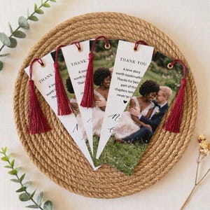 May include: A set of white paper bookmarks featuring a photograph of a couple, the text THANK YOU, and a love story worth bookmarking. Each bookmark has a gold eyelet and a deep red tassel. The design includes a small black heart icon.