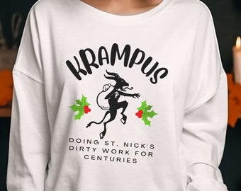 May include: White long-sleeved sweatshirt with a black and white illustration of Krampus, a Christmas demon, with the text "Krampus Doing St. Nick's Dirty Work For Centuries".