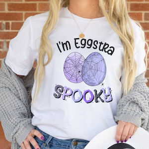 Puede incluir: Camiseta blanca con un diseño de telaraña de araña púrpura y blanca y el texto "I'm Eggstra Spooky".