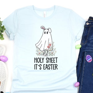 Puede incluir: Camiseta azul claro con un fantasma de dibujos animados con orejas de conejo y flores. El texto "Holy Sheet It's Easter" está impreso en la camiseta.