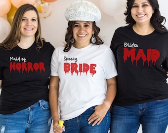 May include: Three friends wearing black and white t-shirts with red dripping text. The text on the shirts reads "Maid of Horror", "Spooky Bride", and "Brides Maid".