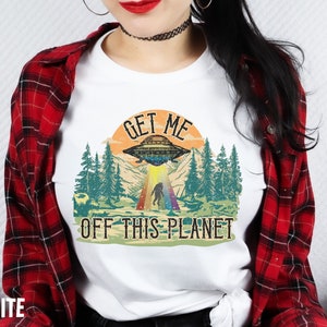 May include: White t-shirt with a retro graphic design featuring a UFO, a Bigfoot, and the text "Get Me Off This Planet".