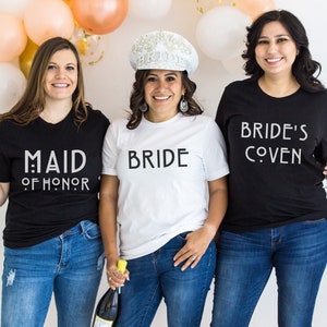 May include: Three people wearing black and white t-shirts with text that reads 'Maid of Honor', 'Bride', and 'Bride's Coven'. The person wearing the 'Bride' shirt is also wearing a white crown.