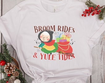 Puede incluir: Camiseta blanca con una ilustración de dibujos animados de una bruja volando en una escoba con una bolsa de regalos. El texto "Broom Rides & Yule Tides" está impreso en la camiseta.