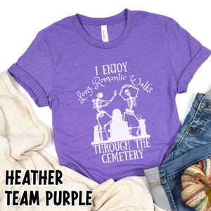 May include: Heather team purple t-shirt with the text "I Enjoy Long Romantic Walks Through The Cemetery" and a graphic of dancing skeletons. The shirt is made of a soft material and has rolled-up sleeves.