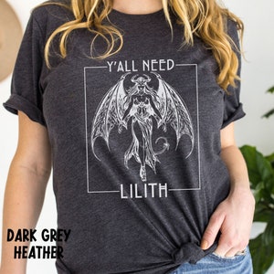 Puede incluir: Camiseta de color gris oscuro jaspeado con un gr&aacute;fico blanco de una mujer demonio con alas y el texto "Y'all need Lilith".