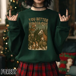 May include: Dark green sweatshirt with the text "YOU BETTER WATCH OUT" above a graphic of forest creatures. The graphic includes a skeleton horse, gnomes, and a wolf. The person in the image is wearing a black choker and a red plaid skirt.