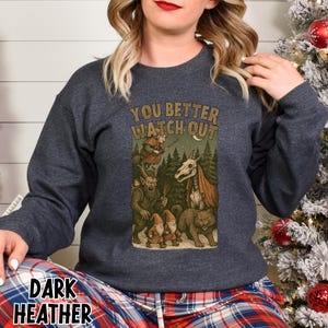 May include: Dark heather gray sweatshirt with a graphic featuring the text "YOU BETTER WATCH OUT" above a forest scene with mythical creatures. The design includes a witch, gnomes, and a skeletal horse-like figure.