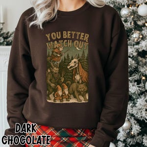 May include: Dark chocolate-colored sweatshirt with the text "YOU BETTER WATCH OUT" above a graphic of forest creatures. The graphic includes a witch, gnomes, a skeletal horse, and a cat. The sweatshirt is a crew neck style.