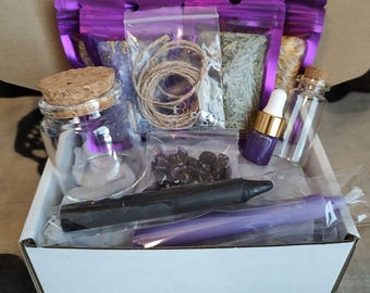 May include: A white box filled with various spiritual items. Includes small glass jars with corks, purple pouches, a black candle, a purple candle, twine, and small bags of crystals and herbs. The box is open, revealing the contents.