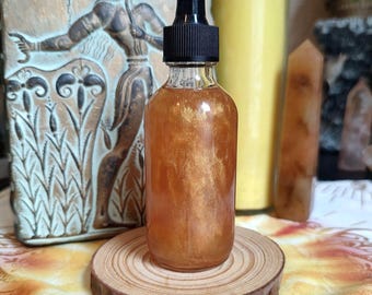 May include: A clear glass bottle with a black dropper top, filled with a shimmering golden liquid. The bottle sits on a small, round wooden slice. The background includes a decorative relief and candles.