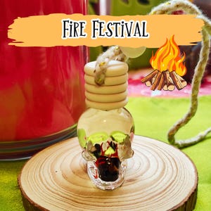 May include: A clear glass skull-shaped air freshener with a wooden top and a rope. The skull has green and red accents. The air freshener sits on a wooden slice. The text "Fire Festival" is above the air freshener.