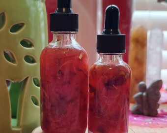 May include: Two glass bottles with dropper tops filled with a pink liquid containing flower petals. The bottles are sitting on a wooden surface.