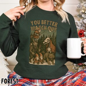 May include: Dark green sweatshirt with the text "YOU BETTER WATCH OUT" above a graphic of forest creatures. The graphic includes a witch, a horned creature, gnomes, a wolf, and a skeleton. The word "FOREST" is at the bottom.