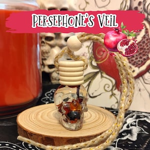 May include: A clear skull-shaped car air freshener with a wooden top and a braided rope. The air freshener contains red and orange liquid and sits on a wooden slice. The image includes a pomegranate and the text "PERSEPHONE'S VEIL".