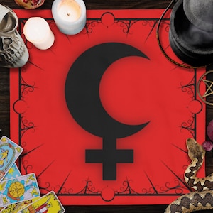 May include: A red cloth with a black crescent moon and cross symbol, surrounded by black floral designs. The cloth is on a wooden surface with tarot cards, candles, a skull, a cauldron, and other occult items.