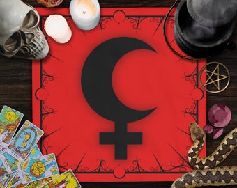 May include: A red cloth with a black crescent moon and cross symbol, surrounded by black floral designs. The cloth is on a wooden surface with tarot cards, candles, a skull, a cauldron, and other occult items.