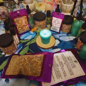 Peut inclure: Une pochette violette remplie d'herbes s&eacute;ch&eacute;es, &eacute;tiquet&eacute;e "Ostara Blessings Simmer Pot Blend". La pochette est entour&eacute;e d'autres pochettes, pots et bouteilles d'herbes et d'huiles, ainsi que d'une bougie turquoise sur un dessous-de-plat en bois. L'arri&egrave;re-plan est un tissu &agrave; motifs floraux.