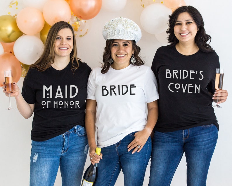 May include: Three people wearing black and white t-shirts with text that reads 'Maid of Honor', 'Bride', and 'Bride's Coven'. The person wearing the 'Bride' shirt is also wearing a white crown.