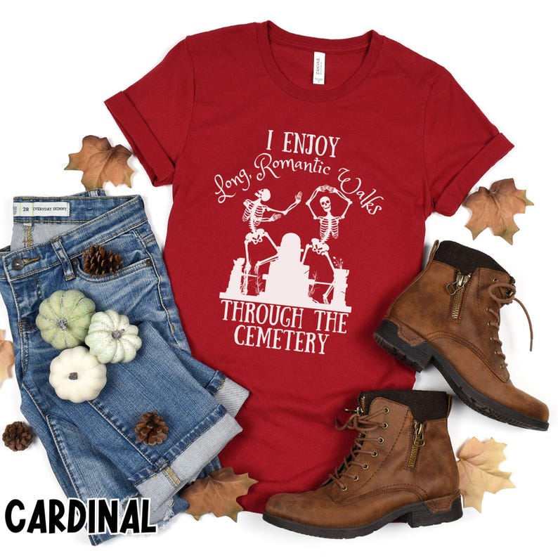 May include: A red t-shirt with the text "I Enjoy Long Romantic Walks Through The Cemetery" and a graphic of dancing skeletons. The image also includes blue denim shorts, brown boots, and decorative leaves.