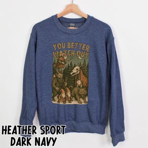 May include: Dark navy blue sweatshirt with a graphic featuring fantasy creatures and the text "YOU BETTER WATCH OUT". The graphic includes a witch, a skeletal horse, gnomes, and a wolf. The sweatshirt is made of heather sport fabric.