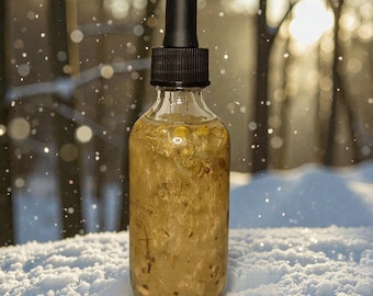 May include: A clear glass bottle with a black dropper top, filled with a golden-colored liquid containing visible plant matter. The bottle is set against a snowy backdrop with a blurred, sunlit forest in the background.
