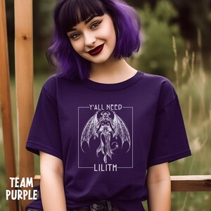 Puede incluir: Camiseta morada con un gr&aacute;fico blanco de un demonio alado con el texto "Y'all need Lilith" en un cuadro blanco.