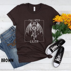 Puede incluir: Una camiseta marr&oacute;n con un gr&aacute;fico blanco de un demonio con alas y el texto "Y'all need Lilith" en un cuadro.