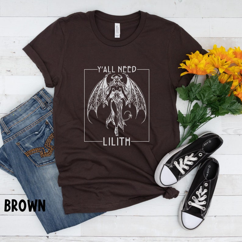 Puede incluir: Una camiseta marr&oacute;n con un gr&aacute;fico blanco de un demonio con alas y el texto "Y'all need Lilith" en un cuadro.