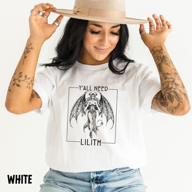 Puede incluir: Camiseta blanca con un gr&aacute;fico en blanco y negro de una mujer demonio con alas y el texto "Y'all need Lilith" en un cuadro negro.