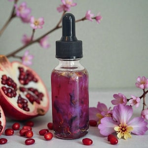 May include: A clear glass bottle with a black dropper filled with a pink liquid and floral elements. The bottle is surrounded by a halved pomegranate, red berries, and pink flowers. The overall aesthetic is natural and botanical.