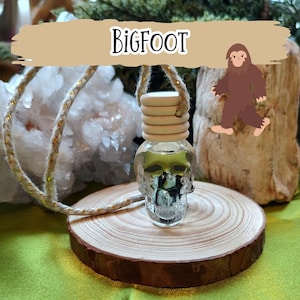 May include: A clear, skull-shaped car air freshener with a wooden top and a Bigfoot graphic. The air freshener hangs from a gold and beige rope, resting on a wooden slice. The word "BIGFOOT" is displayed above the air freshener.
