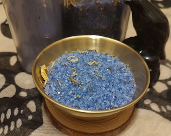 May include: A small, round, brass bowl filled with blue crystals and dried herbs sits on a wooden coaster. A dark blue candle and a glass jar with similar contents are in the background. A black bird figurine is to the right.