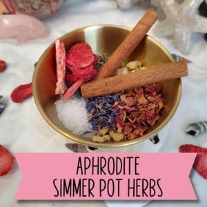 May include: A gold-colored bowl filled with dried strawberries, cinnamon sticks, blue cornflowers, rose petals, and other herbs. The text "APHRODITE SIMMER POT HERBS" is displayed on a pink banner at the bottom of the image.