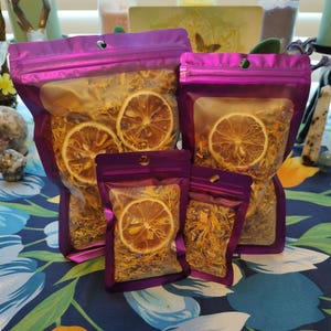 May include: Four purple resealable bags filled with dried lemon slices and other dried herbs. The bags are arranged in a pyramid shape on a blue and green floral patterned surface.