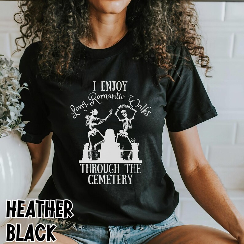 May include: Black t-shirt with white text and graphic. The text reads "I Enjoy Long Romantic Walks Through The Cemetery." The graphic features two skeletons dancing near a tombstone. The words "Heather Black" are at the bottom.
