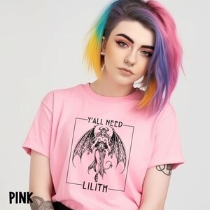 Puede incluir: Camiseta rosa con un gr&aacute;fico en blanco y negro de una mujer con alas y cuernos. El texto "Y'all need Lilith" est&aacute; impreso en la camiseta.