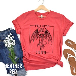 Puede incluir: Camiseta roja de mezcla con un gr&aacute;fico en blanco y negro de una mujer con alas y cola. El texto "Y'all Need Lilith" est&aacute; impreso en la camiseta.