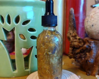 May include: A clear glass bottle with a black dropper top, filled with a golden liquid and botanical elements. The bottle sits on a small, round wooden stand. The background includes a green ceramic object and candles.