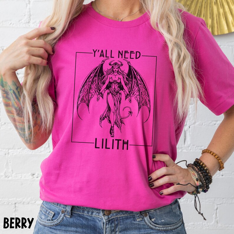 Puede incluir: Una camiseta rosa con un gr&aacute;fico negro de una mujer con alas y el texto "Y'all need Lilith".