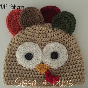 Pode incluir: Chapéu de peru em crochê com detalhes em feltro marrom, vermelho, verde e laranja. O chapéu tem olhos em crochê branco com botões pretos e um bico em crochê amarelo e vermelho. O chapéu tem o texto "Sissa & Bubs" na parte inferior.