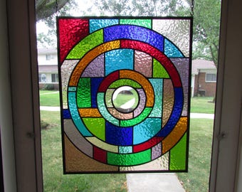 May include: A square stained glass panel featuring a geometric design of concentric circles and rectangular segments in vibrant colours including red, blue, green, orange, and clear textured glass, all held together by dark metal lead lines.