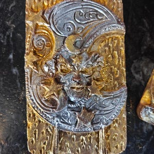 May include: A rectangular, gold-colored decorative piece with a detailed relief design. The design features a crescent moon, stars, and a face. The moon and face are silver-colored, while the background is gold. The piece has a textured surface.