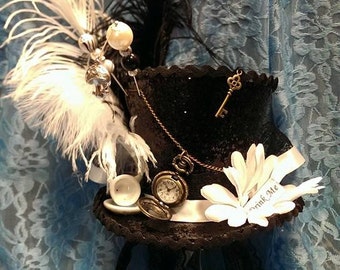 May include: Black top hat with white and black feathers, a pocket watch, and a key charm. The hat has a white ribbon and a flower with the words "Drink Me". A decorative accessory for themed events.