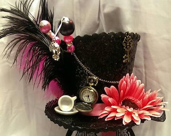 May include: Black top hat with pink and black feathers, a pink flower, and a pocket watch. The hat is decorated with a key charm, beads, and a small teacup. A black lace headband and pink ribbon complete the design.
