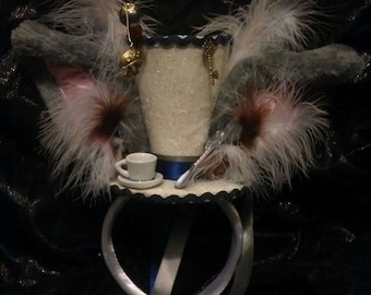 May include: A whimsical, decorative hat with a small teacup and saucer, adorned with white feathers, a gold key, and a pocket watch charm. The hat features a blue ribbon and white ribbons, creating a unique, fantasy-themed accessory.