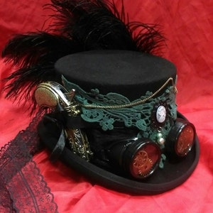 Puede incluir: Un sombrero de copa negro con una banda de encaje verde, una cadena dorada y un pequeño camafeo blanco. El sombrero también tiene una pluma negra, una pistola plateada y dos gafas redondas con engranajes.